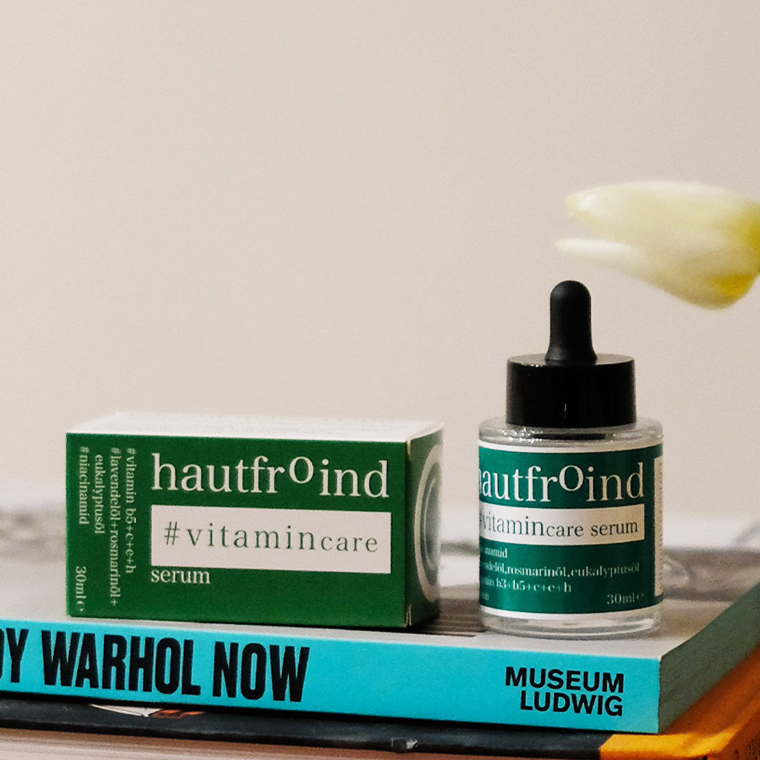 Hautfroind Vitamincare Serum – vitaminreiche Hautpflege und Gesichtspflege mit Niacinamid, Vitamin C & E. Hellt dunkle Flecken auf, fördert die Kollagenproduktion & schützt vor freien Radikalen. Feuchtigkeitsspendende Formel mit Aloe Vera & Hyaluronsäure für strahlende, gesunde Haut. Es ist zu sehen wie die Flasche Hautfroind Vitamincare Serum neben der, auf der Seite liegenden Verpackung, steht. Auf einem Bücherstapel. I Hintergrund ist die Blüte der Tulpe zu sehen. 