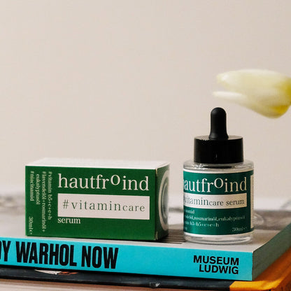 Hautfroind Vitamincare Serum – vitaminreiche Hautpflege und Gesichtspflege mit Niacinamid, Vitamin C & E. Hellt dunkle Flecken auf, fördert die Kollagenproduktion & schützt vor freien Radikalen. Feuchtigkeitsspendende Formel mit Aloe Vera & Hyaluronsäure für strahlende, gesunde Haut. Es ist zu sehen wie die Flasche Hautfroind Vitamincare Serum neben der, auf der Seite liegenden Verpackung, steht. Auf einem Bücherstapel. I Hintergrund ist die Blüte der Tulpe zu sehen. 