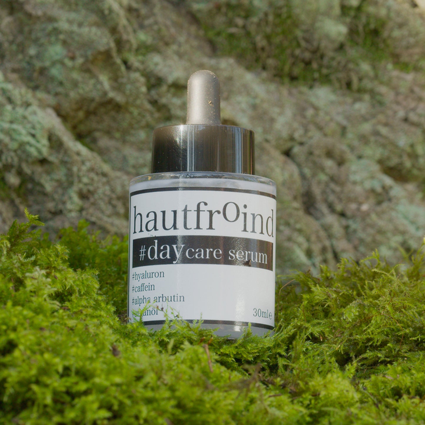 Hautfroind Daycare Serum – feuchtigkeitsspendende Hautpflege für das Gesicht mit Hyaluronsäure, Arganöl & Rosa Damascena Blütenwasser. Stärkt die Hautbarriere, verfeinert den Teint & sorgt für langanhaltende Feuchtigkeit. Leichte, schnell einziehende Gesichtspflege für strahlende Haut. Die Flache mit dem Serum ist vor einer Felswand zu sehen, stehend auf grünem Moos.