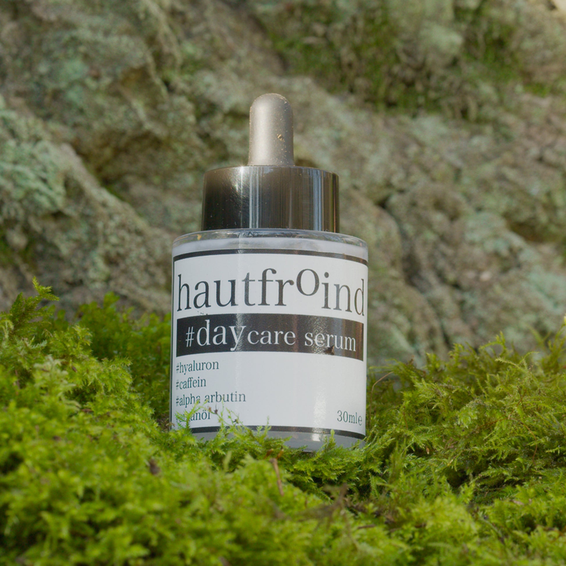 Hautfroind Daycare Serum – feuchtigkeitsspendende Hautpflege für das Gesicht mit Hyaluronsäure, Arganöl & Rosa Damascena Blütenwasser. Stärkt die Hautbarriere, verfeinert den Teint & sorgt für langanhaltende Feuchtigkeit. Leichte, schnell einziehende Gesichtspflege für strahlende Haut. Die Flache mit dem Serum ist vor einer Felswand zu sehen, stehend auf grünem Moos.