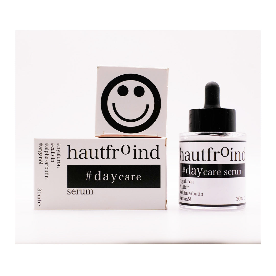 Hautfroind Daycare Serum – feuchtigkeitsspendende Hautpflege für das Gesicht mit Hyaluronsäure, Arganöl & Rosa Damascena Blütenwasser. Stärkt die Hautbarriere, verfeinert den Teint & sorgt für langanhaltende Feuchtigkeit. Leichte, schnell einziehende Gesichtspflege für strahlende Haut. Auf dem Foto ist das Daycare Serum und die Verpackung, liegend, zu sehen. Und die Oberseite der Verpackung mit einem Smiley liegt auf der Verpackung aufgelegt. 