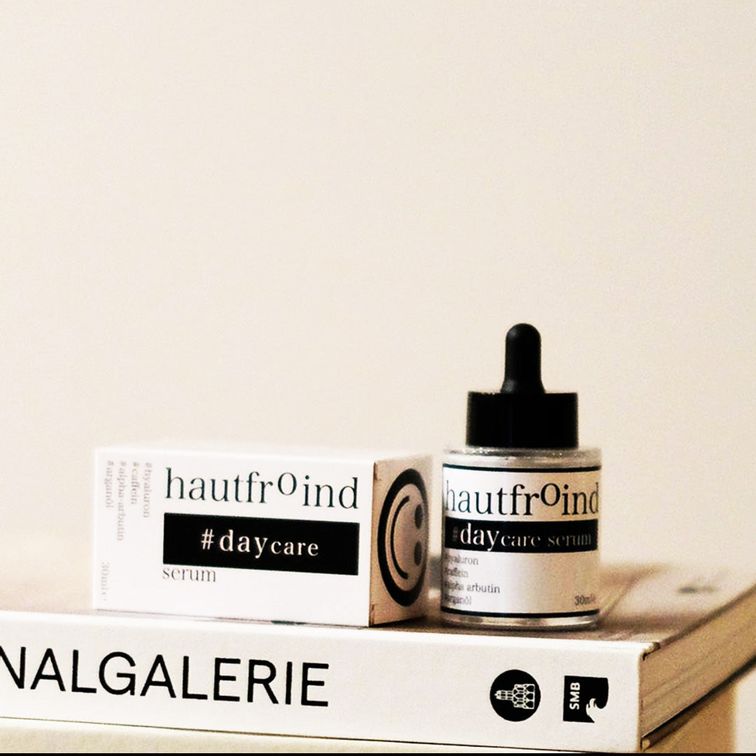 Hautfroind Daycare Serum – feuchtigkeitsspendende Hautpflege für das Gesicht mit Hyaluronsäure, Arganöl & Rosa Damascena Blütenwasser. Stärkt die Hautbarriere, verfeinert den Teint & sorgt für langanhaltende Feuchtigkeit. Leichte, schnell einziehende Gesichtspflege für strahlende Haut. Auf dem Foto ist die Flasche Daycare Serum und die Verpackung zu sehen. Künstlerisch angeordnet auf einem Bücherstapel.