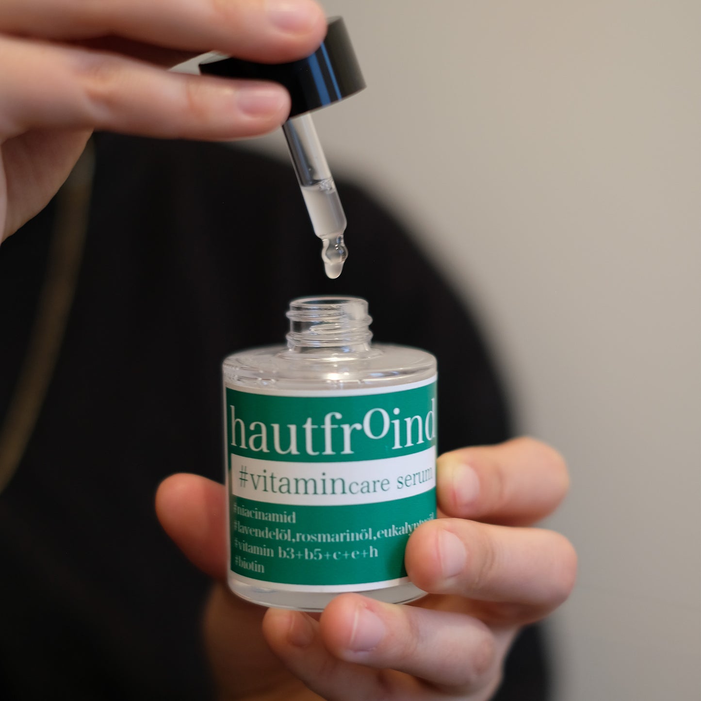 Hautfroind Vitamincare Serum – vitaminreiche Hautpflege und Gesichtspflege mit Niacinamid, Vitamin C & E. Hellt dunkle Flecken auf, fördert die Kollagenproduktion & schützt vor freien Radikalen. Feuchtigkeitsspendende Formel mit Aloe Vera & Hyaluronsäure für strahlende, gesunde Haut. Es ist zu sehen wie zwei Hände die Flasche Hautfroind Vitamincare Serum halten und einen Tropfen des Serums und die Flasche tropft. 