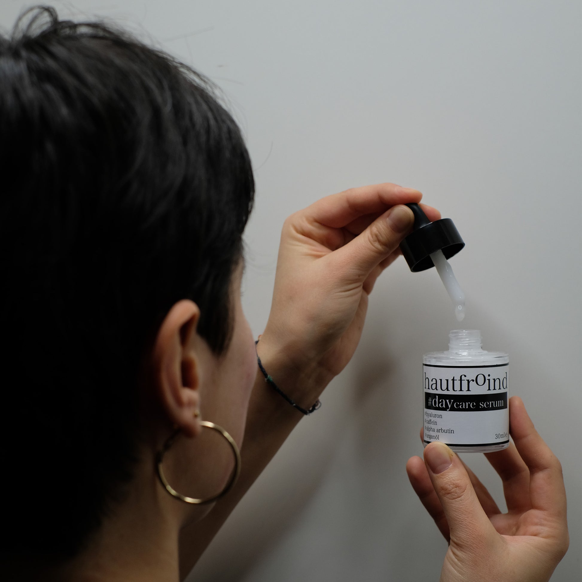 Hautfroind Daycare Serum – feuchtigkeitsspendende Hautpflege für das Gesicht mit Hyaluronsäure, Arganöl & Rosa Damascena Blütenwasser. Stärkt die Hautbarriere, verfeinert den Teint & sorgt für langanhaltende Feuchtigkeit. Leichte, schnell einziehende Gesichtspflege für strahlende Haut. Auf dem Foto ist ein Model zu sehen. Der Hinterkopf der weiblichen Person, wie sie die geöffnete Flasche Daycare Serum vor sich hält.