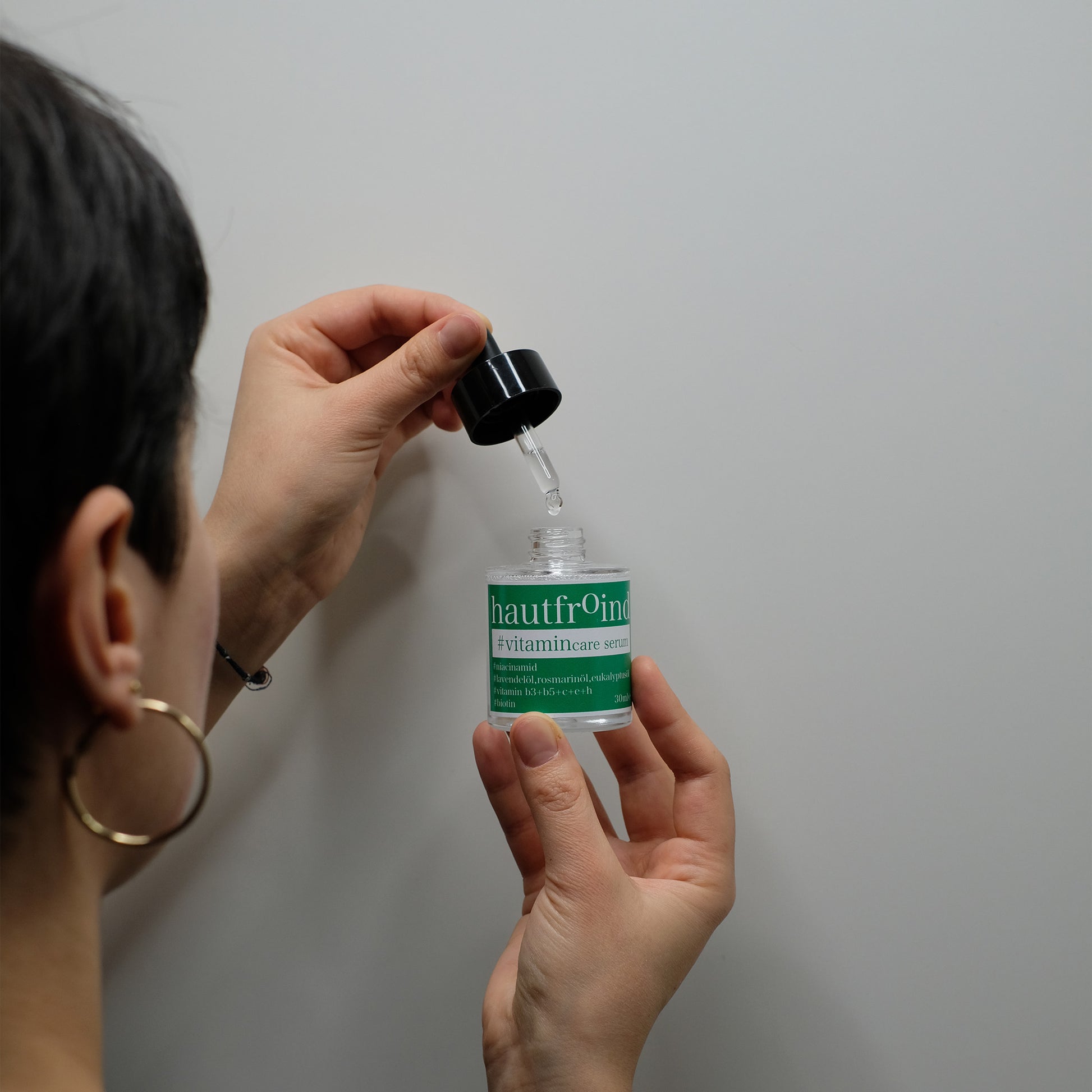 Hautfroind Vitamincare Serum – vitaminreiche Hautpflege und Gesichtspflege mit Niacinamid, Vitamin C & E. Hellt dunkle Flecken auf, fördert die Kollagenproduktion & schützt vor freien Radikalen. Feuchtigkeitsspendende Formel mit Aloe Vera & Hyaluronsäure für strahlende, gesunde Haut. Es ist der Hinterkopf unseres weiblichen Models zu sehen. Sie hat die Flasche Hautfroind Vitamincare Serum geöffnet und tropf das Serum wieder zurück in die Flasche. 